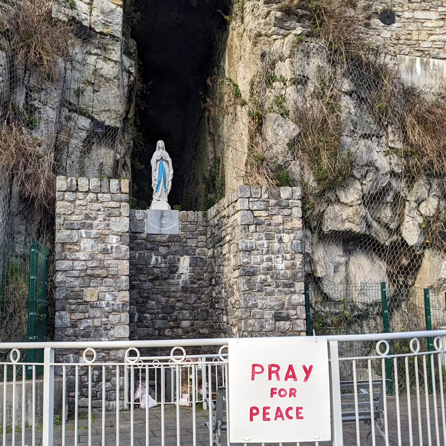 A shrine toOur Lady, set into a rockface. A sign in front says "Pray for Peace" in red lettering.
