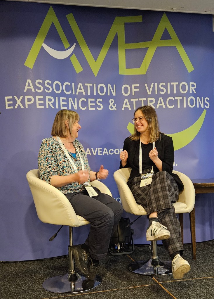 Olivia and artist Sally Booth seated in white chairs engaging in conversation at a conference, with a backdrop displaying the logo of the Association of Visitor Experiences & Attractions.