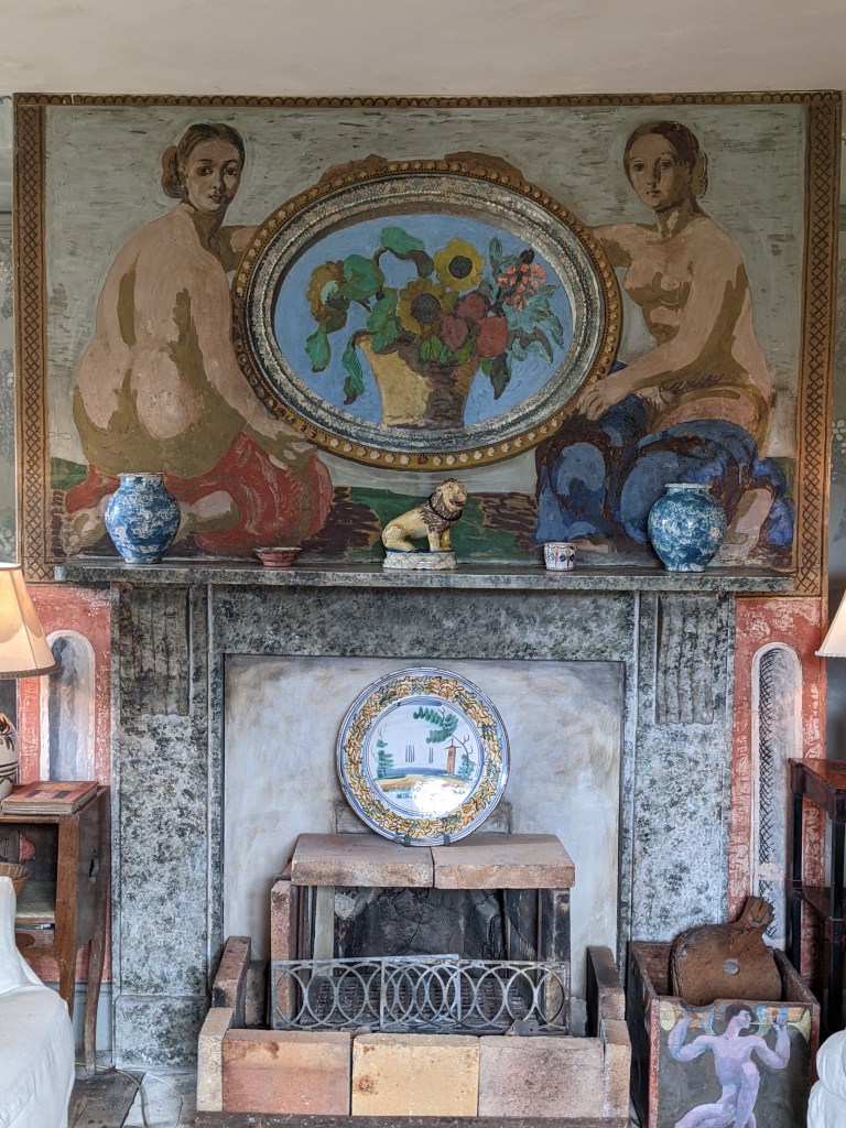 A cozy interior featuring a fireplace with a decorative screen, surrounded by artwork and vases. The wall above the fireplace displays a colorful painting of two seated women and a flower vase. Charleston House.