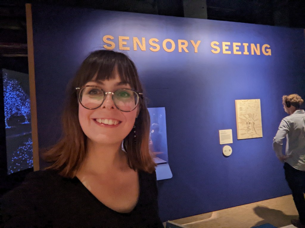 Olivia smiling in front of a cobolt blue exhibition wall labelled 'SENSORY SEEING'.