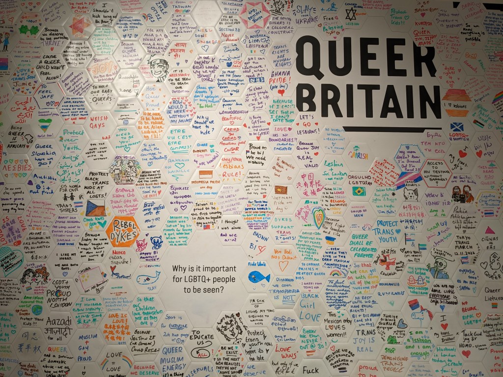 A wall covered with colorful hexagonal notes expressing messages of support and awareness for LGBTQ+ rights, at 'QUEER BRITAIN' in London.