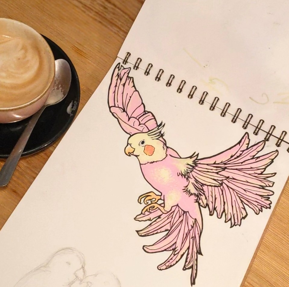 A colored illustration of a flying pink cockatoo in a sketchbook, next to a cup of coffee on a wooden surface.