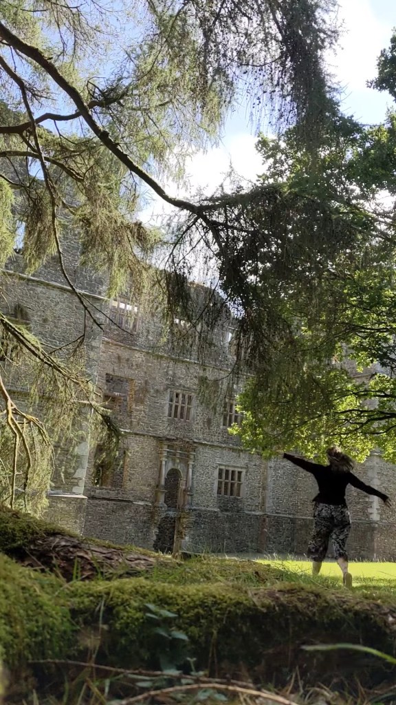 Olivia running with arms raised outdoors in front of an old stone castle, surrounded by trees and lush greenery.